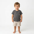 A 3-year-old boy wearing a Palm and Pale charcoal grey Henley t-shirt with a sailor-themed anchor pocket graphic and beige linen shorts against a white background.
