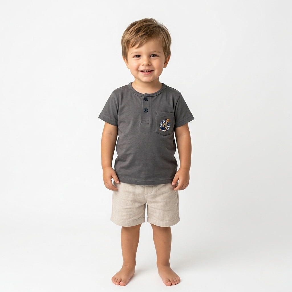 A 3-year-old boy wearing a Palm and Pale charcoal grey Henley t-shirt with a sailor-themed anchor pocket graphic and beige linen shorts against a white background.