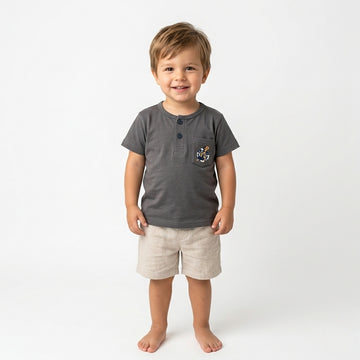 A 3-year-old boy wearing a Palm and Pale charcoal grey Henley t-shirt with a sailor-themed anchor pocket graphic and beige linen shorts against a white background.