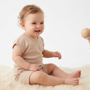 miling baby wearing beige drop-shoulder t-shirt and matching shorts, sitting on soft rug in minimal studio background – summer baby outfit by Palm and Pale