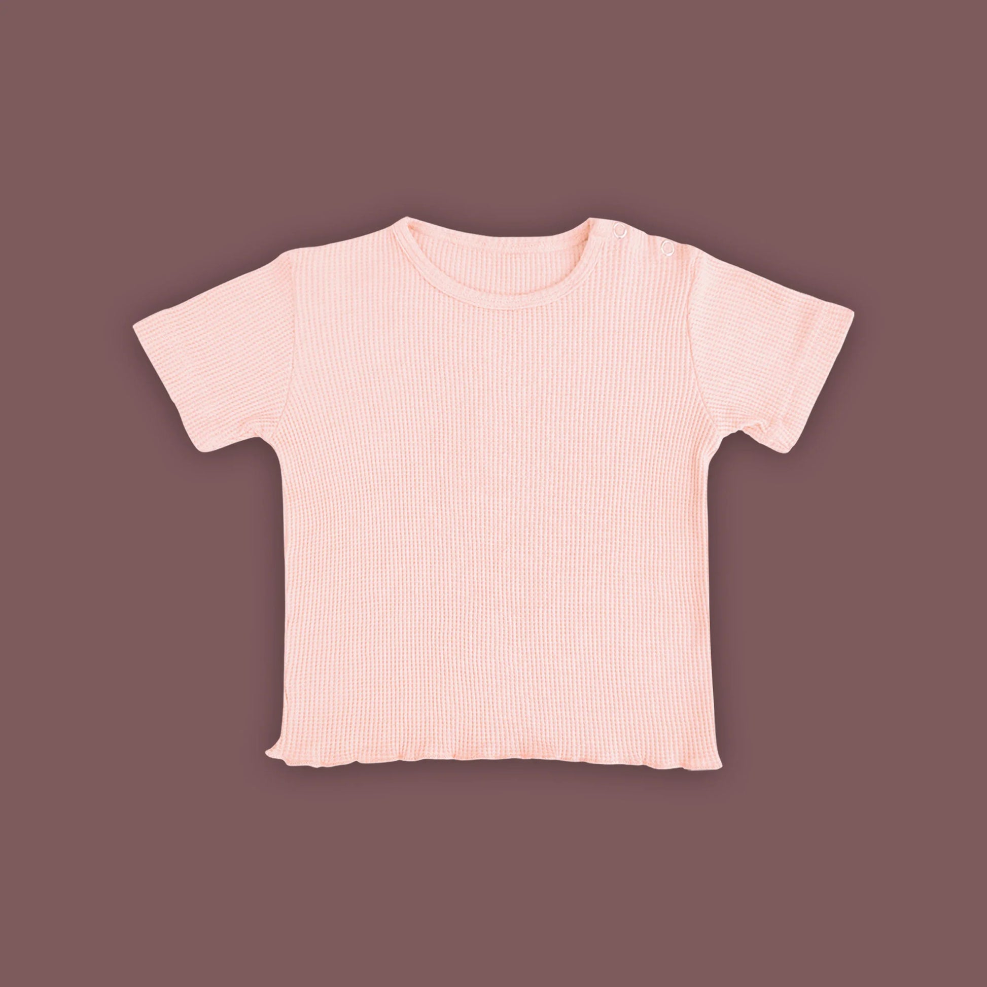 A baby short-sleeved T-shirt in a soft pastel pink color, displayed against a muted brown background. The T-shirt is made from ribbed fabric and features snap buttons on one shoulder for easy dressing. The hem has a subtle wavy finish, adding a delicate touch to the design.