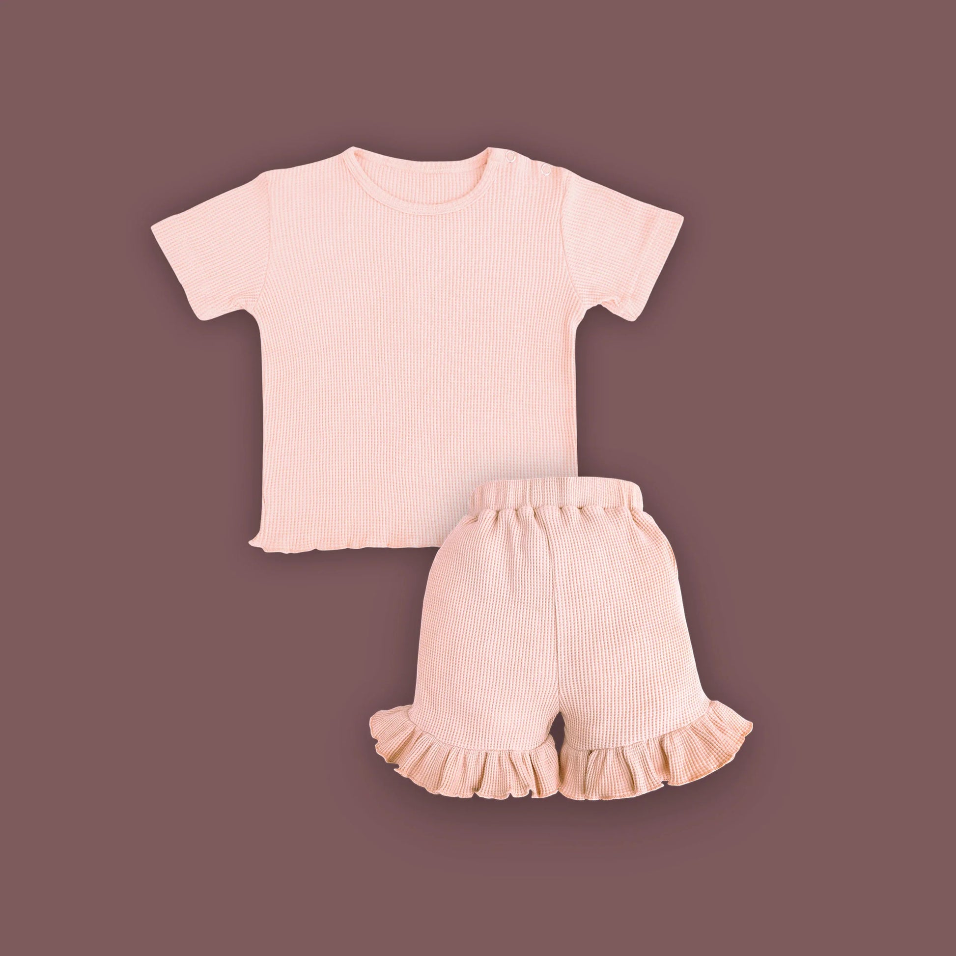 A baby clothing set consisting of a short-sleeved, ribbed T-shirt and ruffled shorts in a soft pastel pink color, displayed on a muted brown background. The T-shirt features snap buttons on one shoulder for easy dressing, and the shorts have an elastic waistband with ruffled hems for a cute and comfortable look.