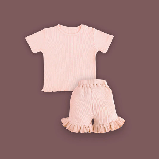 A baby clothing set consisting of a short-sleeved, ribbed T-shirt and ruffled shorts in a soft pastel pink color, displayed on a muted brown background. The T-shirt features snap buttons on one shoulder for easy dressing, and the shorts have an elastic waistband with ruffled hems for a cute and comfortable look.