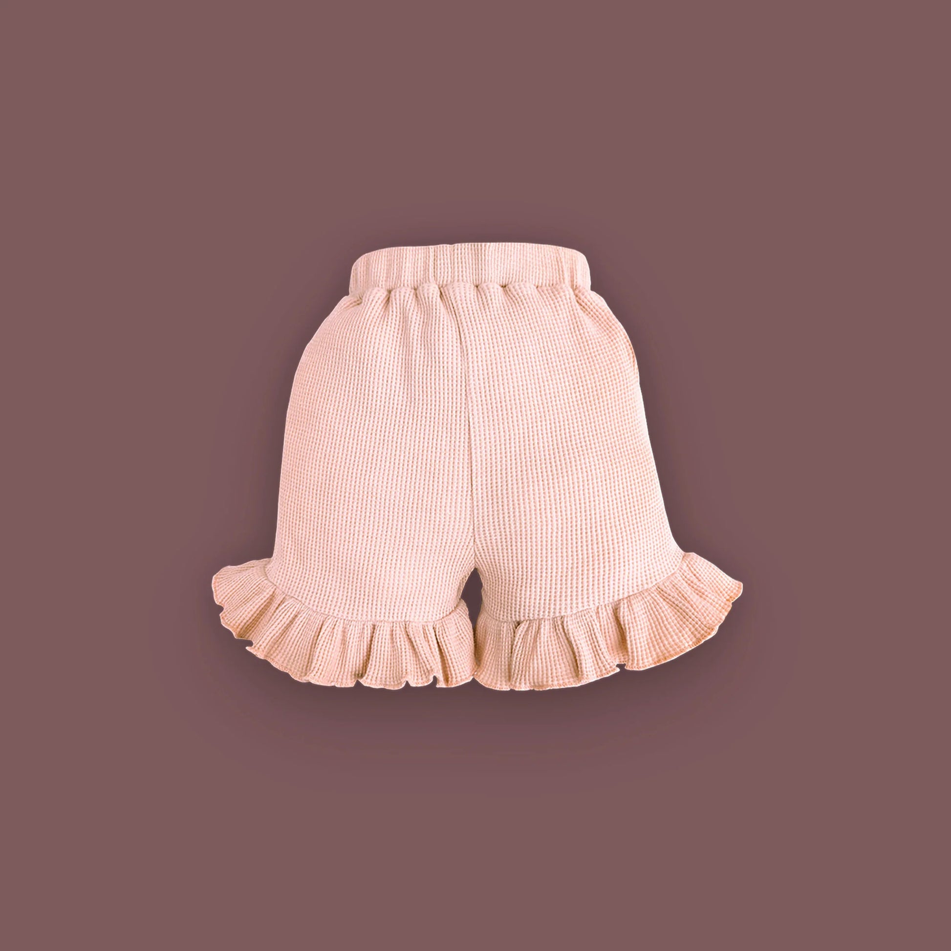 A pair of baby shorts in a soft pastel pink color, displayed against a muted brown background. The shorts are made from ribbed fabric and feature an elastic waistband for comfort. The leg openings are adorned with delicate ruffled hems, adding a cute and stylish touch.