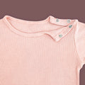 A close-up view of a baby T-shirt in soft pastel pink, highlighting the ribbed fabric texture and shoulder snap button closure. The two metallic snap buttons on one side allow for easy dressing and a comfortable fit. The delicate stitching and gentle stretch of the material are emphasized against a muted brown background.