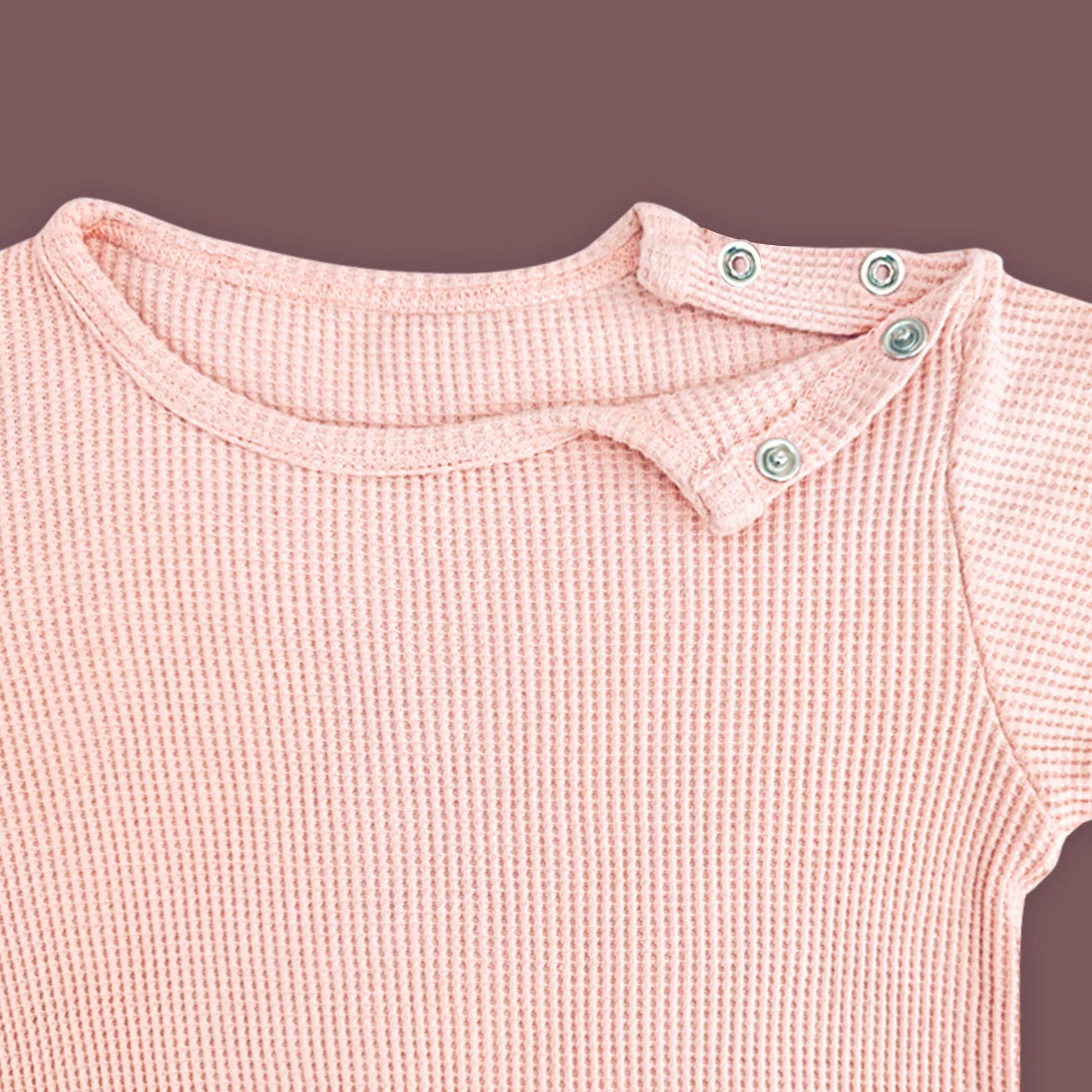 A close-up view of a baby T-shirt in soft pastel pink, highlighting the ribbed fabric texture and shoulder snap button closure. The two metallic snap buttons on one side allow for easy dressing and a comfortable fit. The delicate stitching and gentle stretch of the material are emphasized against a muted brown background.