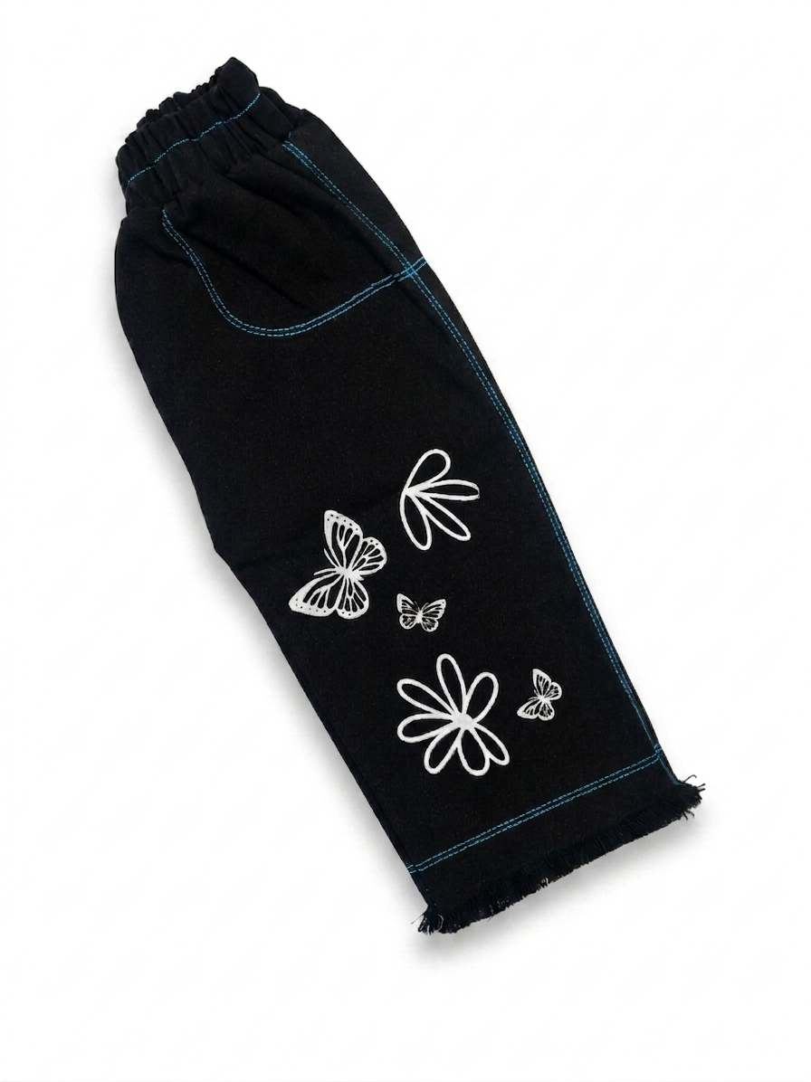 Side view of black girls' denim pants laid flat, showing teal contrast stitching, pocket detail, and butterfly floral print on leg